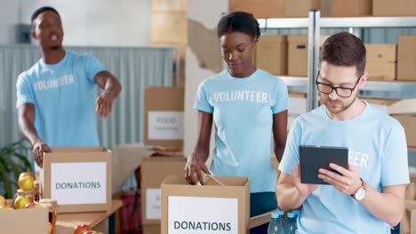 Multiethnic-group-of-volunteers-packing-boxes-with-food-and-clothes-and-typing-on-tablet-in-charity-warehouse