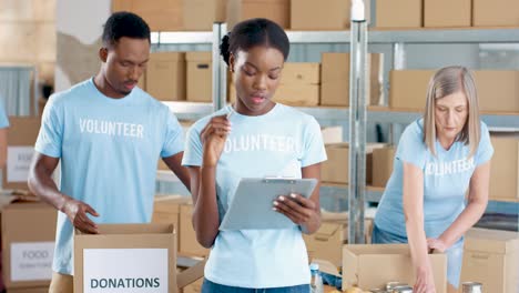 Young-African-American-female-volunteer-writing-and-checking-donations-in-charity-warehouse-while-her-male-coworkers-sorting-donated-stuff-in-boxes-in-the-background