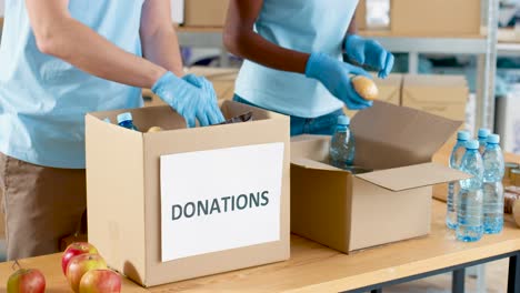 Close-up-view-of-volunteers-hands-packing-box-with-food-and-water-in-charity-warehouse