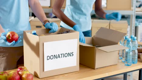 Nahaufnahme-Von-Freiwilligen-übergibt-Verpackungskiste-Mit-Lebensmitteln-Und-Wasser-Im-Lager-Für-Wohltätigkeitsorganisationen