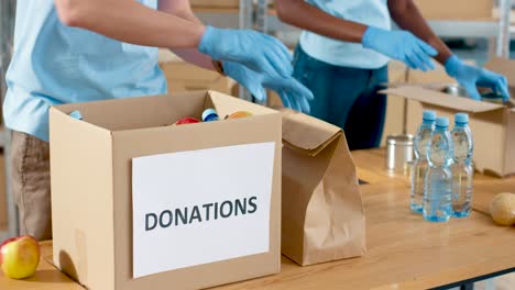 Close-up-view-of-volunteers-hands-packing-box-and-giving-bags-with-food-and-water-to-homeless-people-in-charity-warehouse