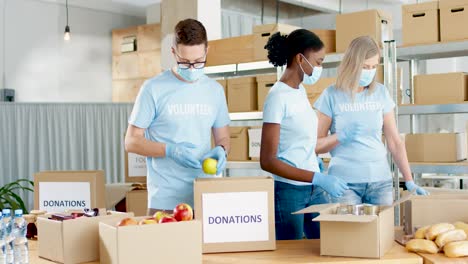 Multiethnic-group-of-volunteers-in-facial-mask-packing-boxes-with-food-and-clothes-in-charity-warehouse