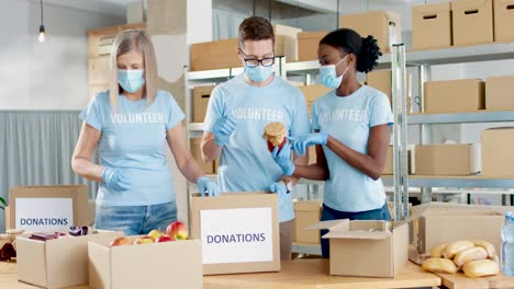 Multiethnic-group-of-volunteers-in-facial-mask-packing-boxes-with-food-in-charity-warehouse