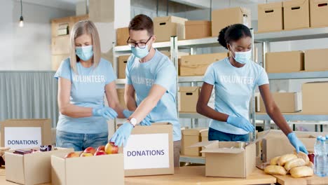 Multiethnic-group-of-volunteers-in-facial-mask-packing-boxes-with-food-and-water-in-charity-warehouse
