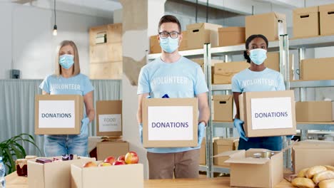 Multiethnic-group-of-volunteers-in-facial-mask-holding-donation-boxes-and-looking-to-the-camera-in-charity-warehouse