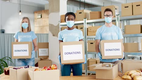 Multiethnic-group-of-volunteers-in-facial-mask-holding-donation-boxes-and-looking-to-the-camera-in-charity-warehouse