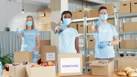 Multi-ethnic-group-of-facial-mask-volunteers-beckoning-with-thumb-up-and-looking-at-camera-in-a-charity-warehouse