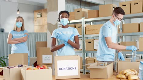 Multiethnic-group-of-volunteers-in-facial-mask-packing-boxes-and-looking-at-the-camera-in-charity-warehouse