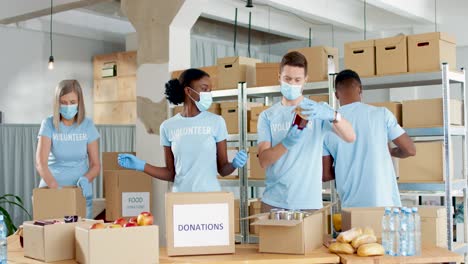 Multiethnische-Gruppe-Von-Freiwilligen-In-Verpackungsboxen-Für-Gesichtsmasken-Mit-Lebensmitteln-Im-Lager-Für-Wohltätige-Zwecke