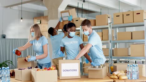 Multiethnic-group-of-volunteers-in-facial-mask-packing-boxes-with-food-in-charity-warehouse