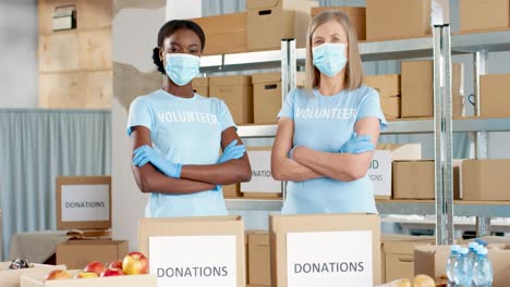African-American-and-caucasian-women-volunteers-in-facial-masks-packing-donation-boxes-and-looking-at-camera-in-charity-warehouse