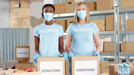 Voluntarias-Afroamericanas-Y-Caucásicas-Con-Máscaras-Faciales-Empacando-Cajas-De-Donación-Y-Mirando-La-Cámara-En-Un-Almacén-De-Caridad