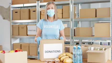 Caucasian-woman-volunteer-in-facial-mask-packing-donation-boxes-and-looking-at-camera-in-charity-warehouse