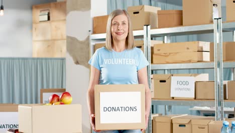 Kaukasische-Freiwillige-Frau,-Die-Spendenboxen-Hält-Und-Kamera-Im-Lager-Für-Wohltätigkeiten-Betrachtet