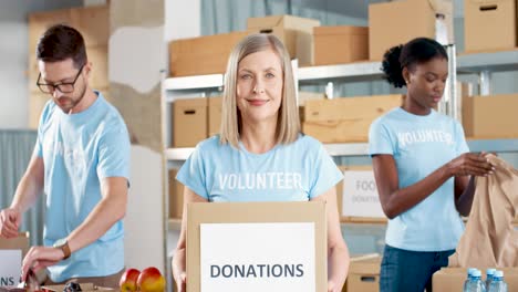 Kaukasische-Freiwillige-Frau,-Die-Spendenboxen-Hält-Und-In-Die-Kamera-Im-Lager-Für-Wohltätigkeit-Schaut,-Während-Ihre-Mitarbeiter-Verpackungsboxen-Bearbeiten