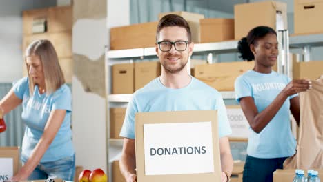 Caucasian-man-volunteer-holding-donation-boxes-and-looking-at-camera-in-charity-warehouse-while-his-coworkers-working-packing-boxes