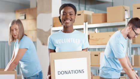 Afrikanisch-amerikanische-Freiwillige,-Die-Spendenboxen-In-Der-Hand-Halten-Und-Im-Lager-Für-Wohltätige-Zwecke-In-Die-Kamera-Schauen,-Während-Ihre-Kollegen-Beim-Packen-Von-Kisten-Arbeiten