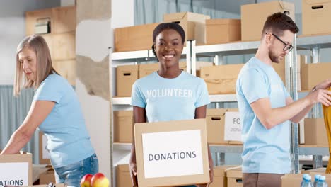 Afrikanisch-amerikanische-Freiwillige,-Die-Spendenboxen-In-Der-Hand-Halten-Und-Im-Lager-Für-Wohltätige-Zwecke-In-Die-Kamera-Schauen,-Während-Ihre-Kollegen-Beim-Packen-Von-Kisten-Arbeiten
