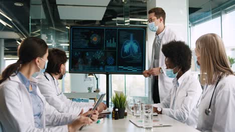 Multiethnisches-Ärzteteam,-Das-Eine-Gesichtsmaske-Trägt-Und-über-Covid-Lungenkrankheiten-Diskutiert,-Während-Sie-Digitale-Bildschirme-Im-Krankenhausbüro-Beobachten