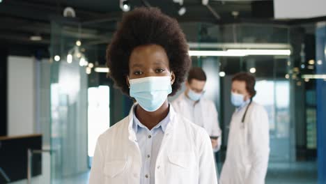 Close-up-view-of-african-american-female-doctor-in-medical-mask-looking-at-camera-standing-in-hospital
