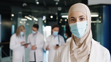 Vista-De-Cerca-De-Una-Doctora-árabe-Con-Máscara-Médica-E-Hiyab-Mirando-La-Cámara-De-Pie-En-El-Hospital