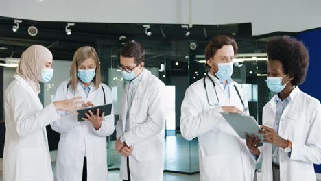 Multiethnic-doctors-team-in-medical-mask-discussing-coronavirus-infection-while-typing-on-tablet-in-hospital