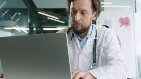 Close-up-view-of-senior-Caucasian-male-doctor-sitting-in-hospital-office-working-and-typing-on-laptop-at-workplace