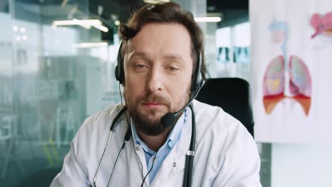 Close-up-view-of-senior-male-doctor-with-headphones-sitting-at-desk-speaking-at-camera-and-giving-medical-advice-about-coronavirus-in-hospital-office