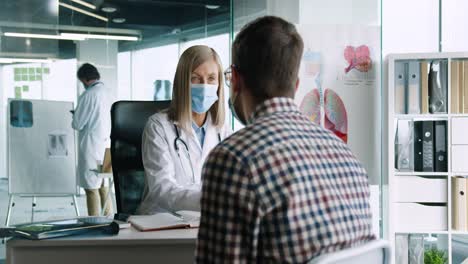 Close-up-view-of-female-doctor-wearing-medical-mask-sitting-at-desk-and-speaking-with-young-patient-while-prescribing-a-treatment