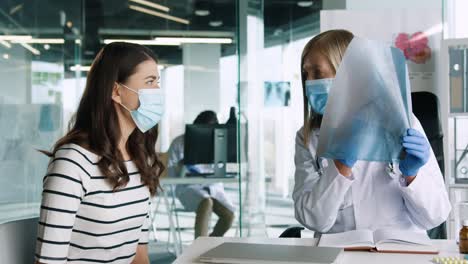 Kaukasische-ärztin-Mit-Medizinischer-Maske,-Die-Am-Schreibtisch-Sitzt-Und-Der-Patientin-In-Der-Medizinischen-Beratung-Die-Lungenkrankheit-Des-Coronavirus-Erklärt