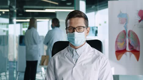 Close-up-view-of-portrait-of-handsome-young-Caucasian-adult-male-doctor-wearing-medical-mask-sitting-in-clinic-at-workplace-looking-at-camera-and-smiling