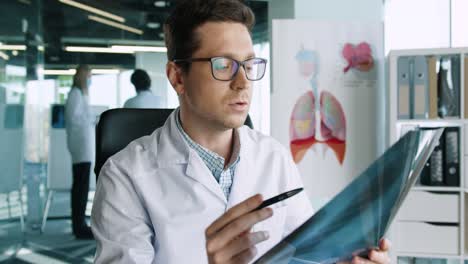 Close-up-view-of-caucasian-male-doctor-in-glasses-sitting-in-clinic-at-workplace-looking-at-camera-and-explining-coronavirus-lung-disease-on-medical-consultation