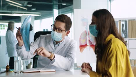 Caucasian-male-doctor-wearing-medical-mask-sitting-at-desk-explaining-coronavirus-lung-disease-to-female-patient-in-medical-consultation