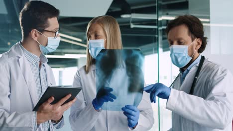 Caucasian-male-and-female-doctors-in-medical-masks-speaking-and-discussing-Xray-scan-and-typing-on-tablet-device-in-hospital-office