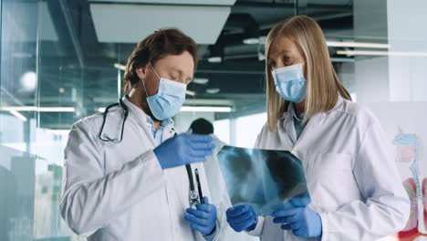 Caucasian-male-and-female-doctors-in-medical-masks-speaking-and-discussing-Xray-scan-in-hospital-office