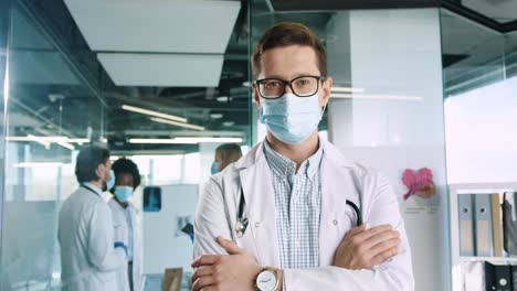 Kaukasischer-Männlicher-Arzt-Mit-Brille-Und-Medizinischer-Maske,-Der-Im-Krankenhausbüro-In-Die-Kamera-Schaut