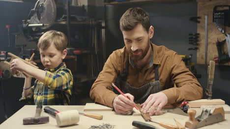 Cheerful-young-Caucasian-carpenter-working-at-desk-in-workshop-and-his-little-son-playing-next-to-him