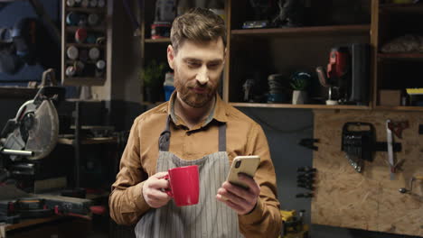 Caucasian-young-male-carpenter-in-apron-resting-in-workshop,-sipping-hot-drink-and-using-smartphone