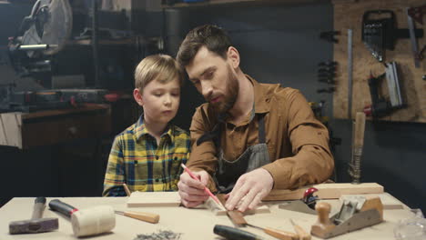 Young-Caucasian-carpenter-teaching-how-to-work-with-wood-his-small-son-in-workshop