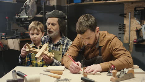 Old-man-carpenter-sitting-at-desk-in-workshop-with-her-son-and-cute-little-grandson