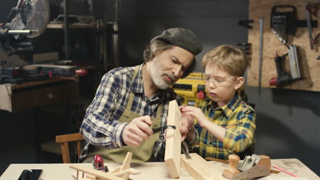 Caucasian-senior-man-carpenter-teaching-his-small-cute-grandson-how-to-work-with-wood-in-workshop-at-desk