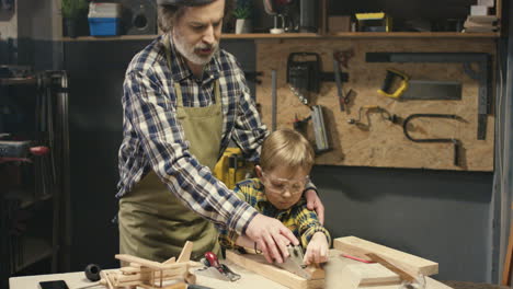 Caucasian-old-carpenter-with-gray-beard-teaching-his-little-grandson-to-work-with-hardwood-in-workshop