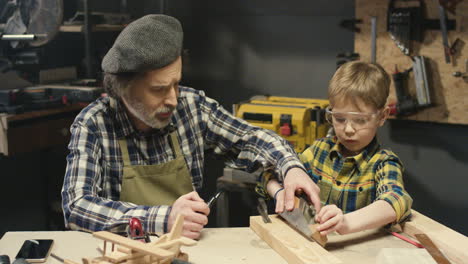 Caucasian-old-carpenter-with-gray-beard-teaching-his-little-grandson-to-work-with-hardwood-in-workshop