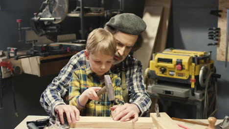 Caucasian-grandfather-carpenter-teaching-small-cute-grandson-to-spike-with-hammer-in-workshop
