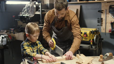 Caucasian-carpenter-man-teaching-his-little-son-to-work-with-hardwood-and-sawing-timber-in-workshop