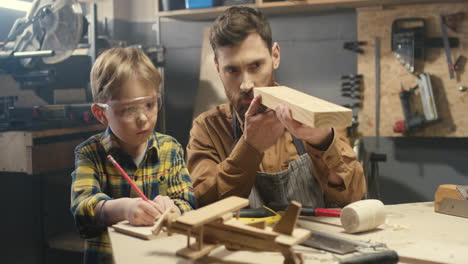 Cheerful-Caucasian-young-father-teaching-small-boy-to-work-with-timbers-in-workshop