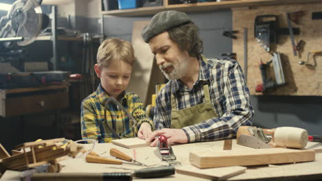 Caucasian-carpenter-man-teaching-his-little-son-to-work-with-hardwood-and-sawing-timber-in-workshop