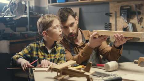 Cheerful-young-Caucasian-carpenter-working-at-desk-in-workshop-while-his-small-son-is-playing-next-to-him
