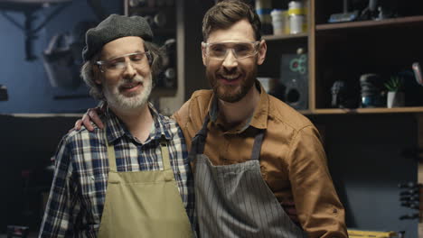 Portrait-shot-of-two-cheerful-father-and-son-in-goggles-hugging-and-looking-at-camera-in-workshop