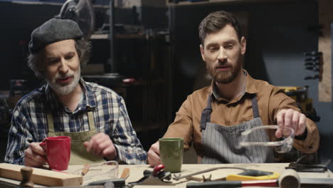Portrait-shot-of-two-cheerful-father-and-son-carpenters-talking-and-drinking-coffee-in-workshop
